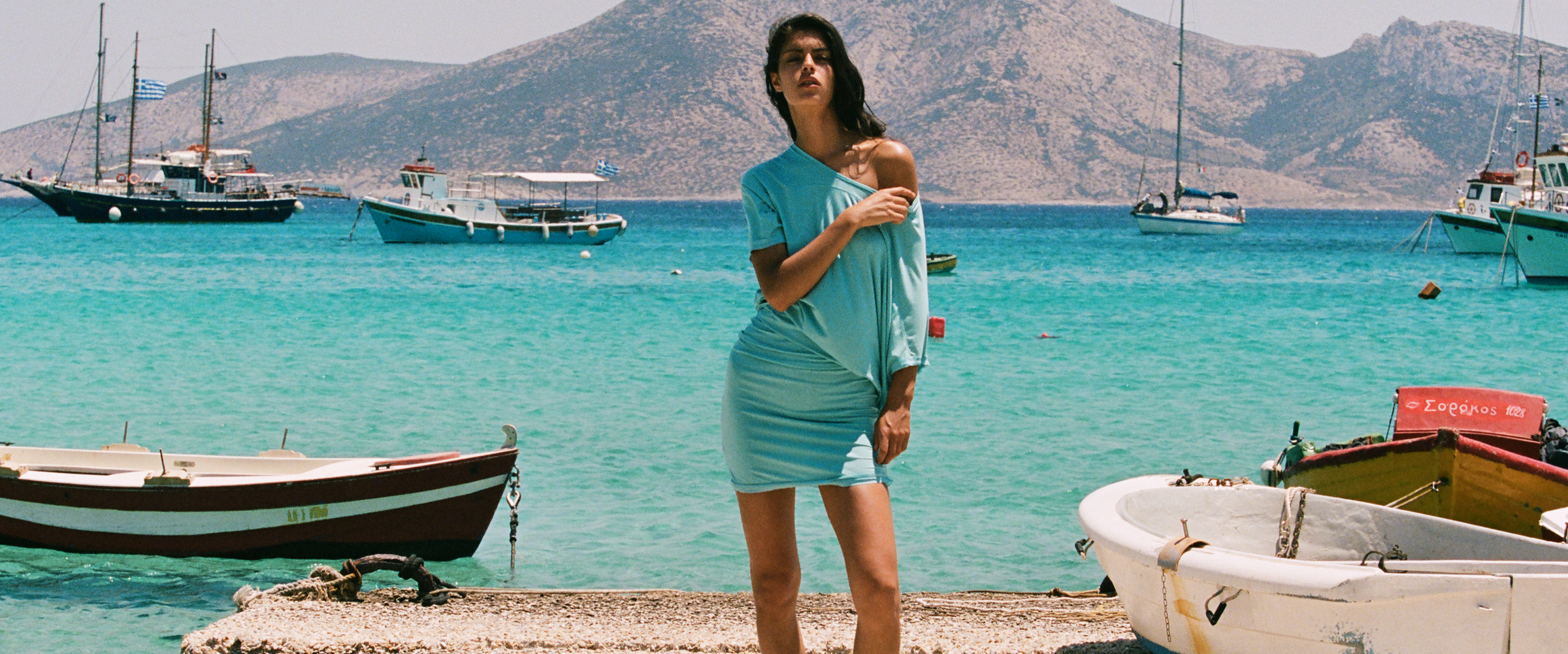 A woman wearing a light blue t-shirt and matching mini skirt stands on a rocky shoreline with turquoise water, boats, and mountains in the background.