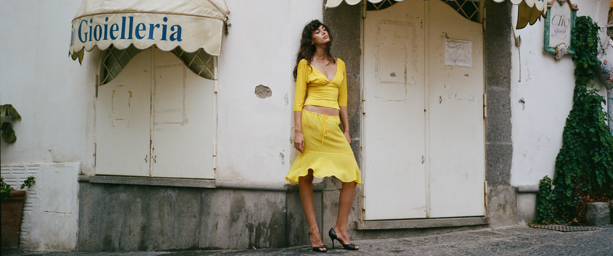 A woman stands outdoors against a weathered white wall wearing a bright yellow two-piece outfit consisting of a long-sleeve crop top and a matching midi skirt.