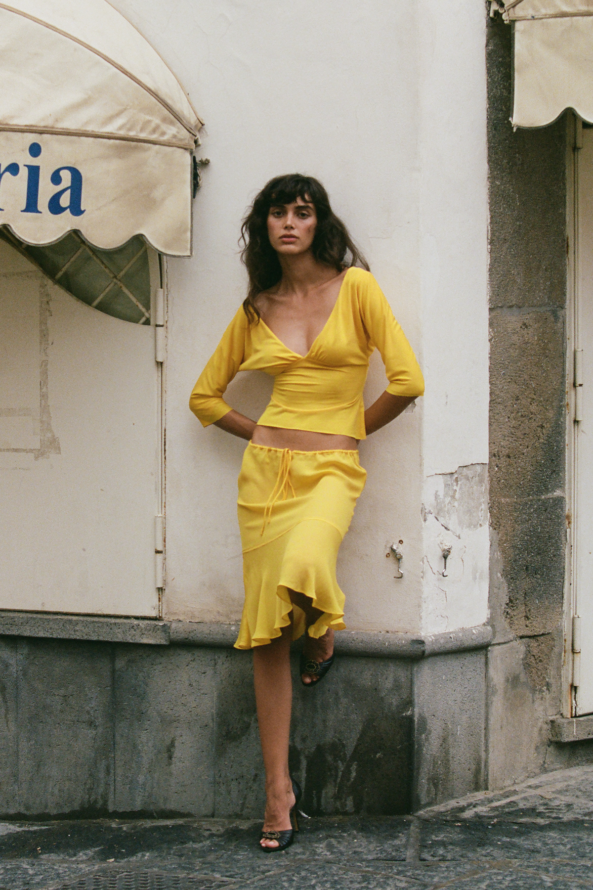 A woman stands outdoors against a white building wall wearing a bright yellow two-piece outfit consisting of a long-sleeve cropped top and a matching low-rise midi skirt.