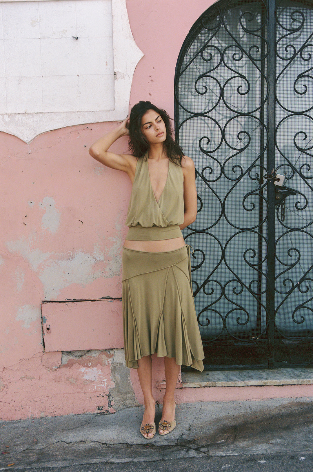 Model in olive Sharni halter wrap top with V-neck and matching midi skirt, posing against a pink wall.