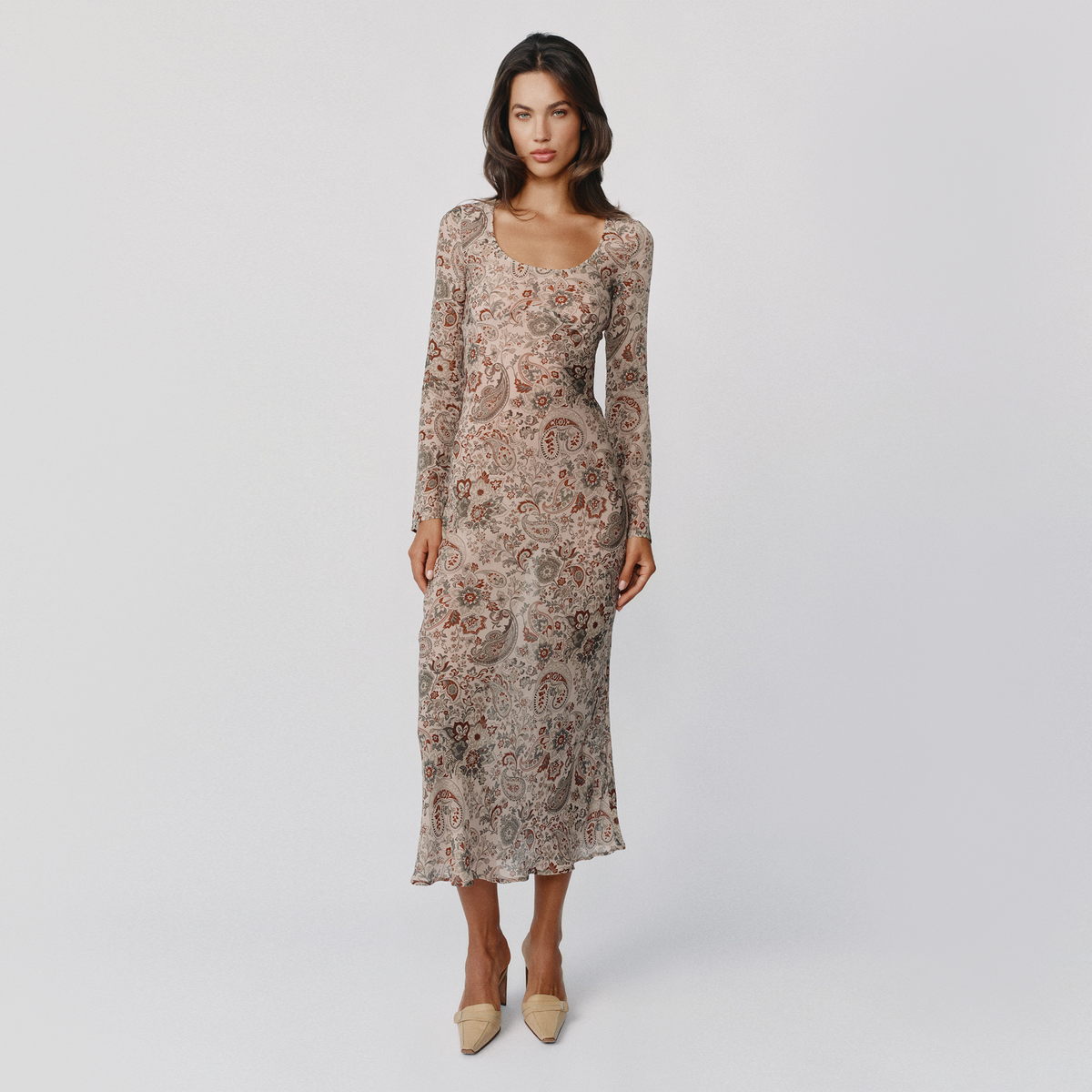 A woman modeling a long-sleeve midi dress with a beige, brown, and rust paisley pattern, paired with beige pointed-toe heels, against a light gray studio background.
