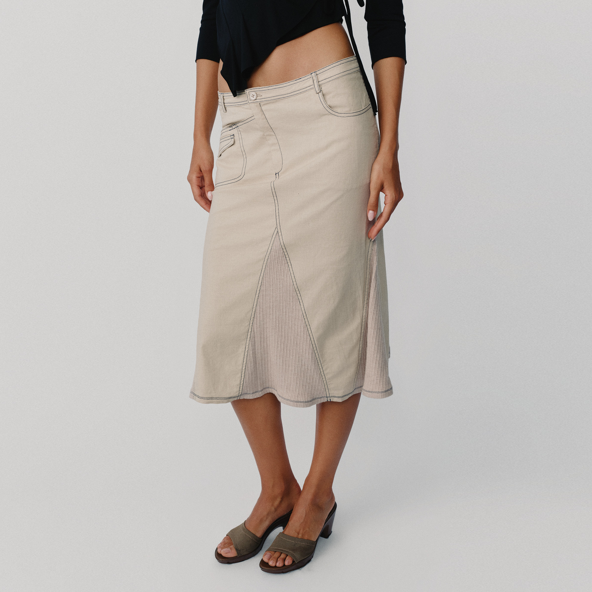 A model wearing a light beige wrap-style skirt with ribbed knit side panels and front pockets, paired with a black cropped top and taupe slide sandals, against a light gray studio background.