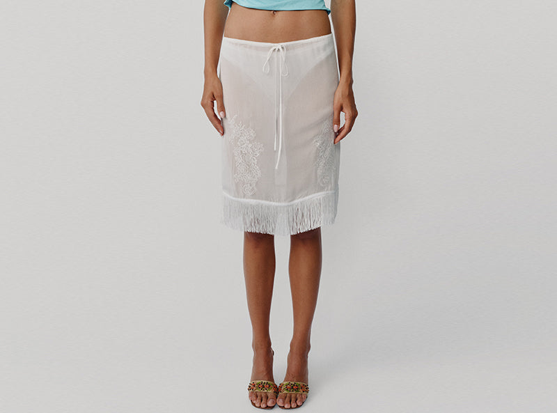 A model wearing a white sheer lace beach cover-up skirt with a drawstring waist and fringe hem, paired with leopard-print flat sandals against a light gray studio background.