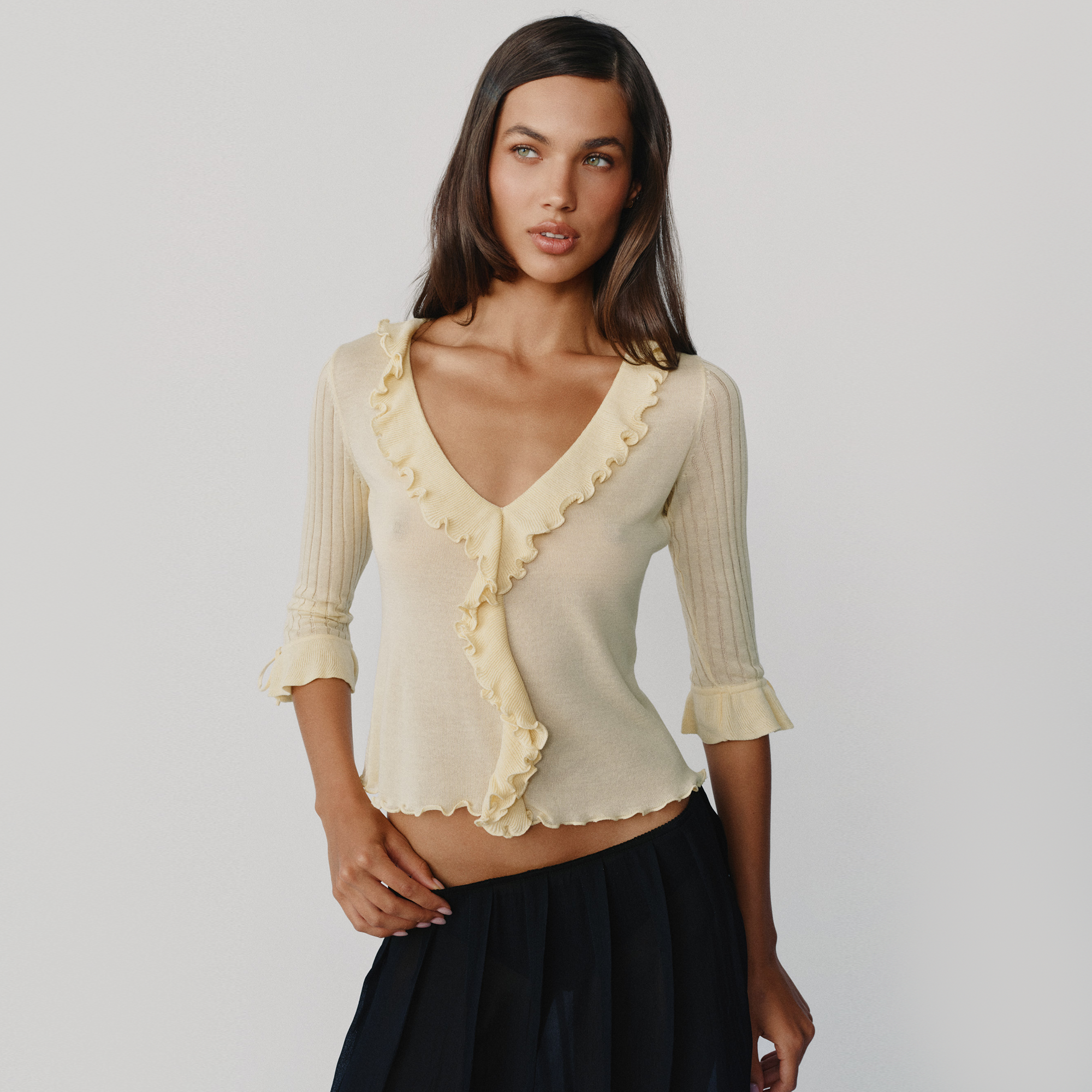 A woman modeling a pale cream ribbed knit top with ruffle trim and three-quarter sleeves, paired with a dark skirt, against a light gray studio background.