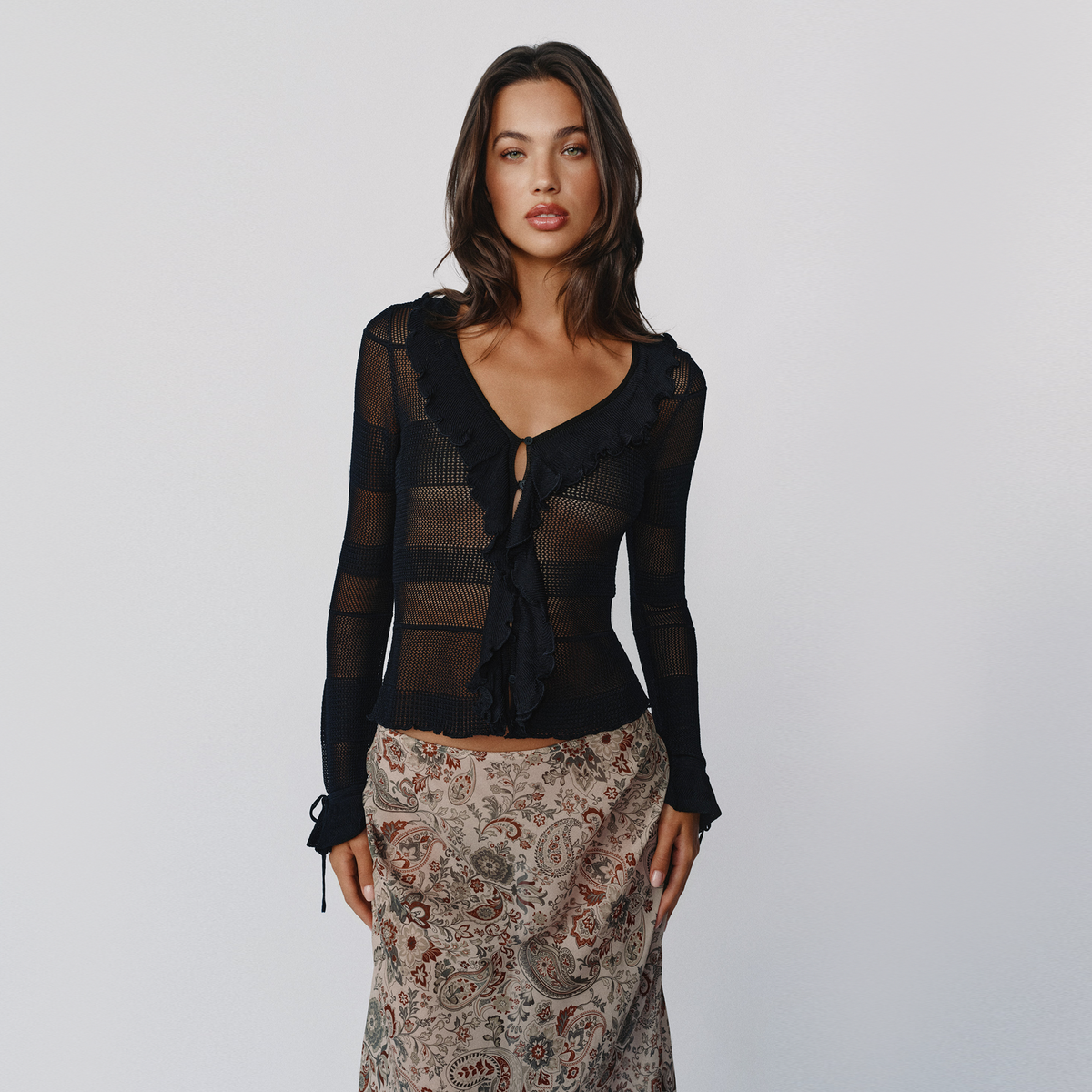 A woman modeling a sheer black long-sleeve top with ruffle trim and a tie-front detail, paired with a beige paisley skirt, against a light gray studio background.