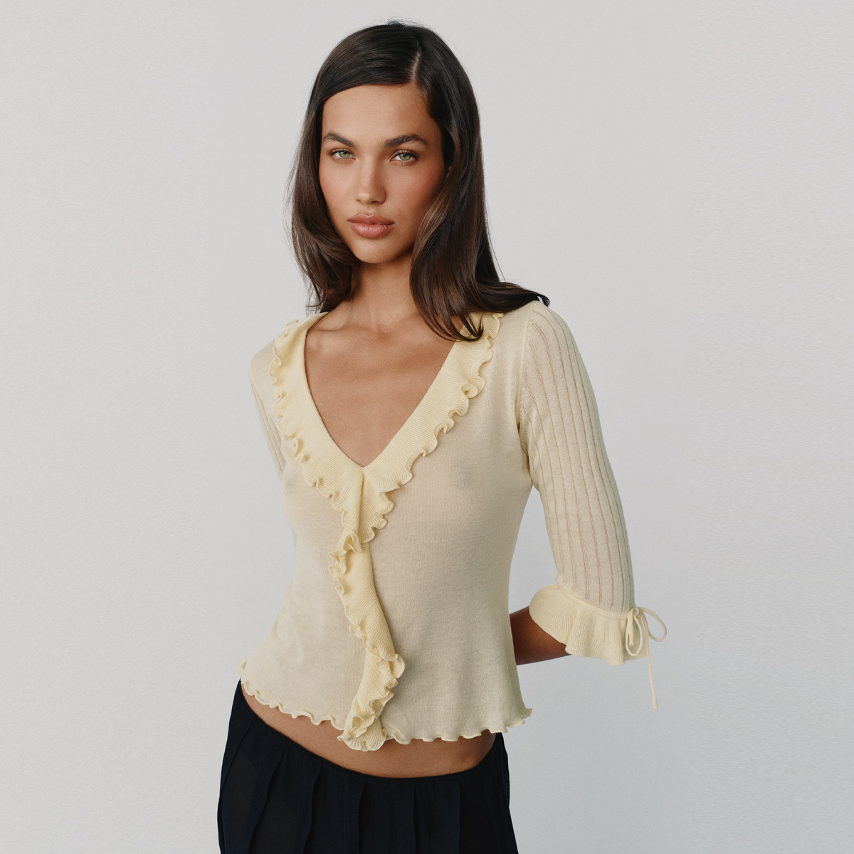 A woman modeling a pale yellow-colored ribbed knit top with ruffle trim and tie details on the sleeves, paired with dark navy bottoms, against a light gray studio background.