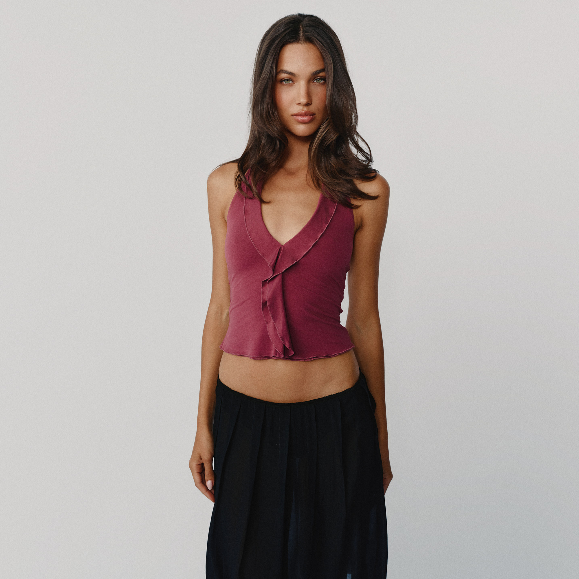A woman modeling a sleeveless crop top in dusty burgundy, paired with black skirt  against a plain light gray studio background.