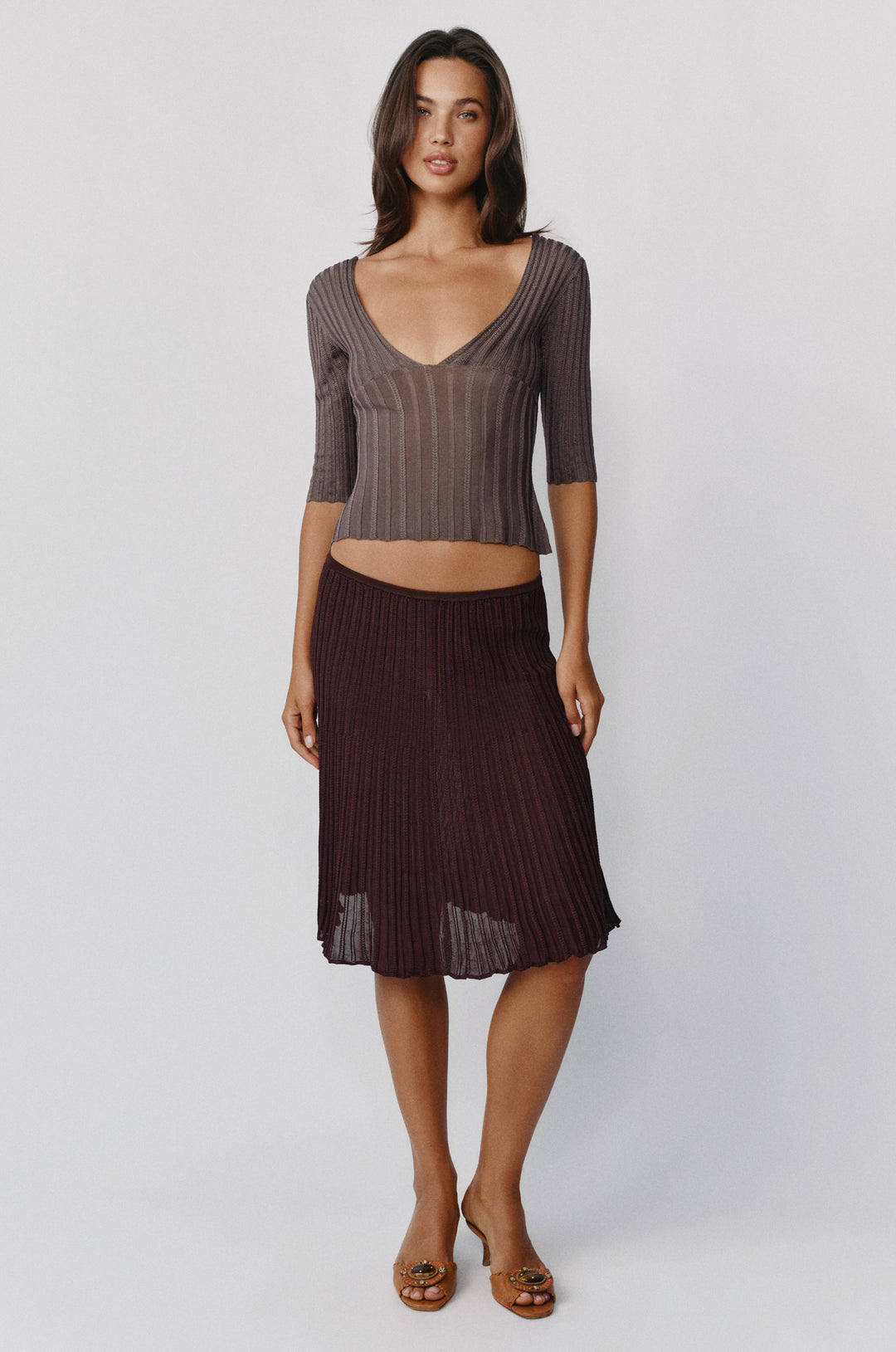Model wearing a smoke-colored Veda ribbed-knit top with a V-neckline and half sleeves paired with a pleated midi skirt.
